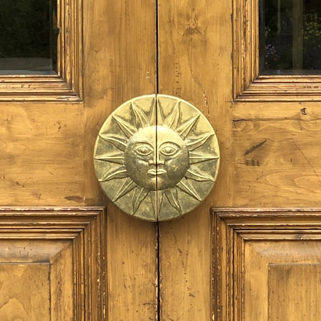 A wooden door features a central, ornate metallic doorknob in the shape of a sun with a face. The wood has a natural golden brown finish with visible grain patterns. The sun motif is detailed with rays extending outward, and the door is symmetrically paneled.