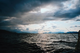Dark, dramatic clouds contrast with a serene body of water, creating a split between the turbulent sky and the calm sea. Mountains line the horizon under a subtle glow from the sun. A lone fishing rod is visible on the right, suggesting a peaceful fishing activity.