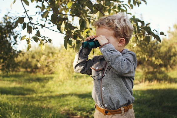 A young child with curly blonde hair wears a grey button-down shirt and holds binoculars to their eyes, looking towards the distance. They are standing outdoors in a lush green setting with trees and sunlight filtering through the leaves.