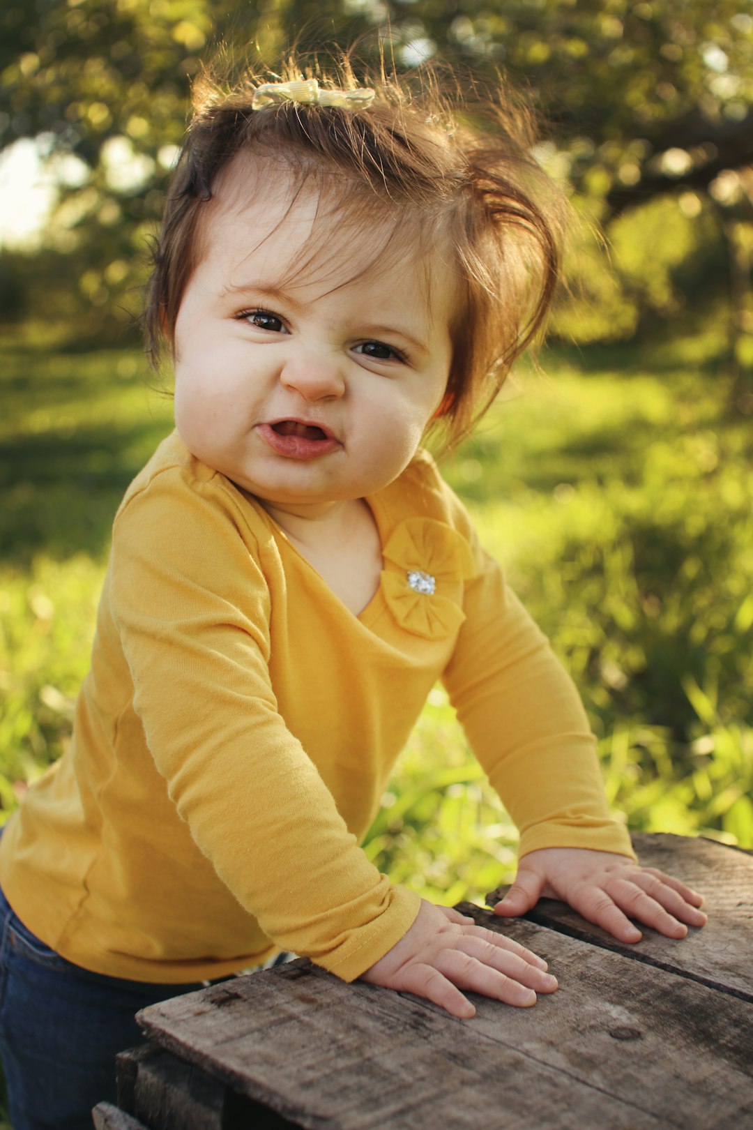Young toddler girl wearing yellow shirt with red hair standing against a wooden crate in apple orchard at golden hour.