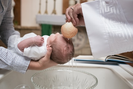 A baby is being held over a baptismal font while water is being poured over its head with a scallop shell. The scene takes place indoors, likely in a church, with hands in ceremonial clothing visible.