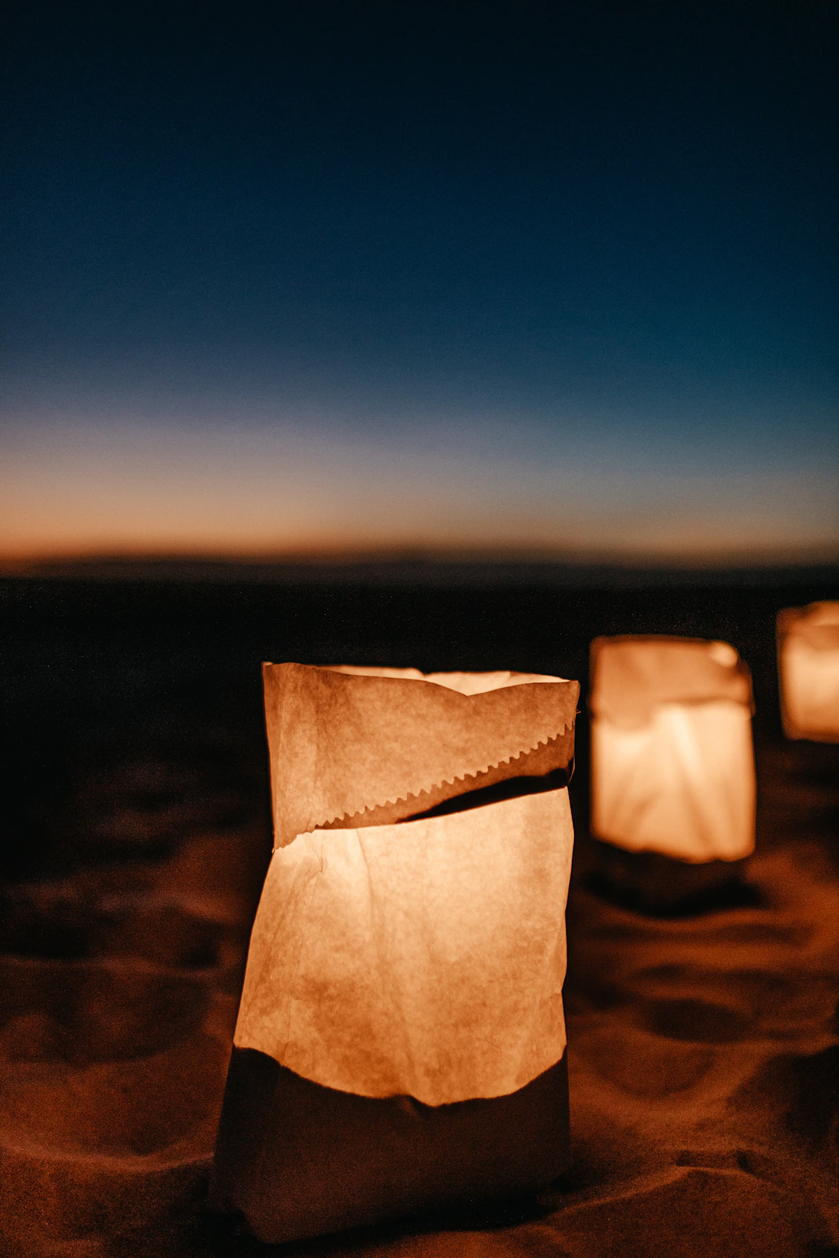 Paper bag lanterns with candles inside glowing along a low wall at dusk