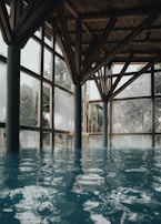 A serene indoor pool with clear blue water, surrounded by large glass windows that let in natural light. The wooden ceiling features intricate beams, creating a warm and rustic atmosphere. Tall trees are visible outside through the windows, adding a touch of nature to the tranquil setting.