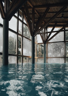 A serene indoor pool with clear blue water, surrounded by large glass windows that let in natural light. The wooden ceiling features intricate beams, creating a warm and rustic atmosphere. Tall trees are visible outside through the windows, adding a touch of nature to the tranquil setting.