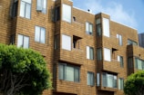 A modern apartment building features geometric architectural design with a series of jutting window frames. The exterior is clad in a warm, tan-colored material resembling wooden shingles. Several windows are slightly open, suggesting a breezy day. Trees with lush green foliage are visible in the foreground, adding a touch of nature to the urban setting.