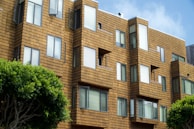 A modern apartment building features geometric architectural design with a series of jutting window frames. The exterior is clad in a warm, tan-colored material resembling wooden shingles. Several windows are slightly open, suggesting a breezy day. Trees with lush green foliage are visible in the foreground, adding a touch of nature to the urban setting.