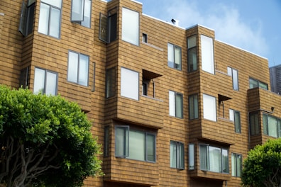 A modern apartment building features geometric architectural design with a series of jutting window frames. The exterior is clad in a warm, tan-colored material resembling wooden shingles. Several windows are slightly open, suggesting a breezy day. Trees with lush green foliage are visible in the foreground, adding a touch of nature to the urban setting.
