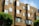A modern apartment building features geometric architectural design with a series of jutting window frames. The exterior is clad in a warm, tan-colored material resembling wooden shingles. Several windows are slightly open, suggesting a breezy day. Trees with lush green foliage are visible in the foreground, adding a touch of nature to the urban setting.