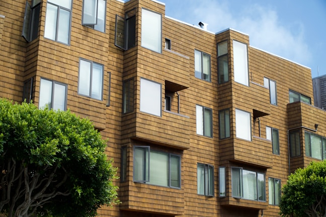 A modern apartment building features geometric architectural design with a series of jutting window frames. The exterior is clad in a warm, tan-colored material resembling wooden shingles. Several windows are slightly open, suggesting a breezy day. Trees with lush green foliage are visible in the foreground, adding a touch of nature to the urban setting.