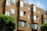 A modern apartment building features geometric architectural design with a series of jutting window frames. The exterior is clad in a warm, tan-colored material resembling wooden shingles. Several windows are slightly open, suggesting a breezy day. Trees with lush green foliage are visible in the foreground, adding a touch of nature to the urban setting.
