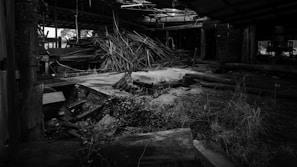 An abandoned industrial space filled with scattered and piled wooden planks. The setting appears deteriorated, with broken structures and overgrown weeds. There are beams and metal debris strewn about, with sunlight filtering through the roof's gaps.