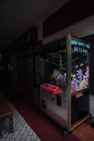 An arcade claw machine filled with various plush toys is situated in a dimly lit hallway. The machine's front panel has a bright red control area with joysticks and buttons, and the enclosure has a city skyline design on the top. The hallway features a combination of dark and light tiled flooring, and there is a wooden bench nearby.
