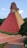 A red pyramid-like structure with a stepped design resembling ancient Mesoamerican architecture. The structure has intricate carvings and designs at the top, with a staircase leading up. It's surrounded by greenery and a paved courtyard.