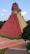 A red pyramid-like structure with a stepped design resembling ancient Mesoamerican architecture. The structure has intricate carvings and designs at the top, with a staircase leading up. It's surrounded by greenery and a paved courtyard.