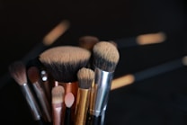 A collection of various makeup brushes with different shapes and sizes is shown, set against a dark background. The bristles vary in color, from beige to dark brown, and the handles are metallic in hues of silver, gold, and rose gold.