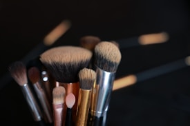 A collection of various makeup brushes with different shapes and sizes is shown, set against a dark background. The bristles vary in color, from beige to dark brown, and the handles are metallic in hues of silver, gold, and rose gold.