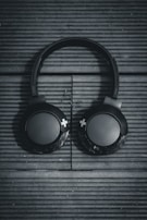 Black over-ear headphones rest on a textured, wooden surface with horizontal lines running across. The design is minimalistic, featuring large circular ear pads with plus symbols on them. The lighting casts a moody shadow, enhancing the sleekness of the headphones.