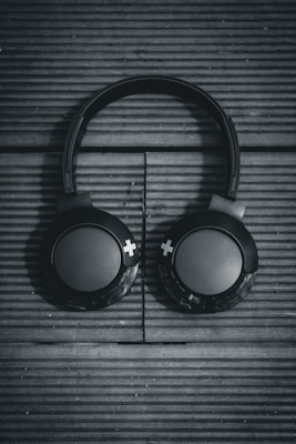 Black over-ear headphones rest on a textured, wooden surface with horizontal lines running across. The design is minimalistic, featuring large circular ear pads with plus symbols on them. The lighting casts a moody shadow, enhancing the sleekness of the headphones.