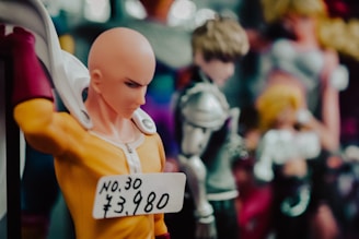 A close-up of a figurine of a bald character in a yellow outfit with red gloves, featuring a price tag reading 'No.30 ¥3,980'. Blurred in the background are other figurines in various poses.