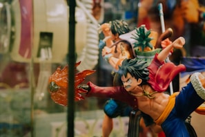 A dynamic action figure depicting a stylized character with spiky hair, wearing a red jacket and blue pants. The figure is posed in an intense action stance, with one arm extended outward holding a fiery effect. Surrounding it are other similar figures, including one holding a pineapple, suggesting a collection of figurines from a themed series.