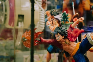 A dynamic action figure depicting a stylized character with spiky hair, wearing a red jacket and blue pants. The figure is posed in an intense action stance, with one arm extended outward holding a fiery effect. Surrounding it are other similar figures, including one holding a pineapple, suggesting a collection of figurines from a themed series.