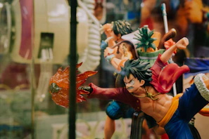 A dynamic action figure depicting a stylized character with spiky hair, wearing a red jacket and blue pants. The figure is posed in an intense action stance, with one arm extended outward holding a fiery effect. Surrounding it are other similar figures, including one holding a pineapple, suggesting a collection of figurines from a themed series.