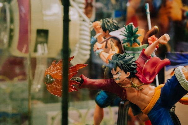 A dynamic action figure depicting a stylized character with spiky hair, wearing a red jacket and blue pants. The figure is posed in an intense action stance, with one arm extended outward holding a fiery effect. Surrounding it are other similar figures, including one holding a pineapple, suggesting a collection of figurines from a themed series.
