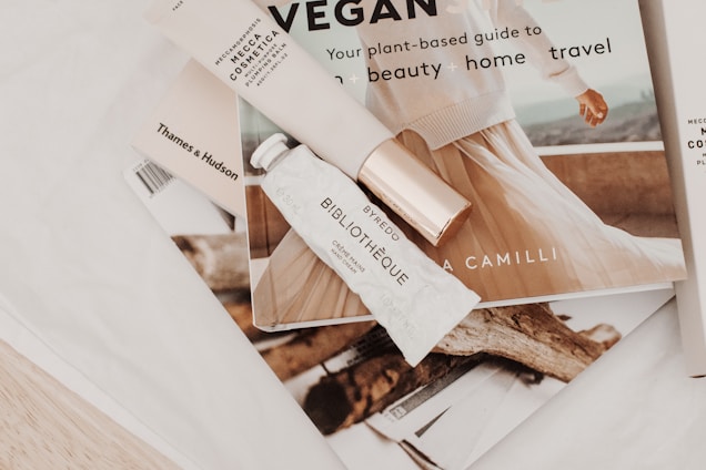 A soft, aesthetically pleasing arrangement featuring a few beauty and lifestyle items. A tube labeled 'Bibliothèque' by Byredo and a 'Mecca Cosmetica' item are resting on an open book or magazine with an elegant image of a person. Other books, including one by Thames & Hudson, are partially visible. The overall scene conveys a sense of sophistication and tranquility.