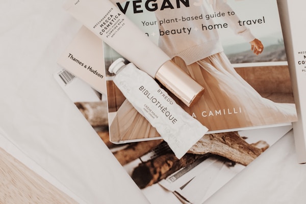 A soft, aesthetically pleasing arrangement featuring a few beauty and lifestyle items. A tube labeled 'Biblioth&egrave;que' by Byredo and a 'Mecca Cosmetica' item are resting on an open book or magazine with an elegant image of a person. Other books, including one by Thames & Hudson, are partially visible. The overall scene conveys a sense of sophistication and tranquility.