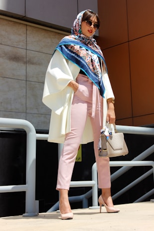 A person is wearing a stylish outfit with a colorful scarf, white coat, pink pants, and sunglasses. They are holding a textured handbag and standing confidently in a bright, outdoor setting.