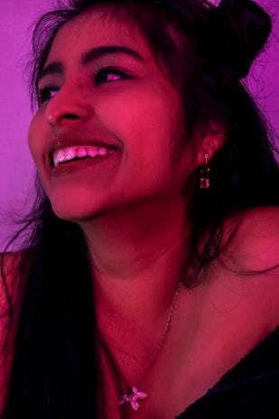 A happy young woman wearing Femiora jewelry, smiling softly in a dimly lit room with shadow pink accents.