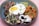 A colorful dish featuring a sunny-side-up egg with a bright yellow yolk, surrounded by grilled meats, seasoned sweet potatoes, mixed cabbage salad, and garnished with edible flowers and microgreens. The dish is arranged in a deep blue bowl placed on a light pink surface.