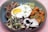 A colorful dish featuring a sunny-side-up egg with a bright yellow yolk, surrounded by grilled meats, seasoned sweet potatoes, mixed cabbage salad, and garnished with edible flowers and microgreens. The dish is arranged in a deep blue bowl placed on a light pink surface.