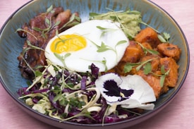 A colorful dish featuring a sunny-side-up egg with a bright yellow yolk, surrounded by grilled meats, seasoned sweet potatoes, mixed cabbage salad, and garnished with edible flowers and microgreens. The dish is arranged in a deep blue bowl placed on a light pink surface.