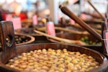 Various wooden barrels filled with different kinds of olives are arranged in a market setting. The olives are submerged in brine, and some barrels contain wooden ladles. A few barrels in the background are blurred, suggesting a busy and vibrant marketplace environment.