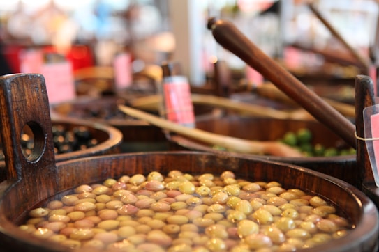Various wooden barrels filled with different kinds of olives are arranged in a market setting. The olives are submerged in brine, and some barrels contain wooden ladles. A few barrels in the background are blurred, suggesting a busy and vibrant marketplace environment.