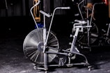 A stationary exercise bike with large fan blades is situated in a dimly lit gym environment. The bike is black with metallic accents and has dual handlebars and a comfortable saddle. Other gym equipment can be seen vaguely in the background, while a person appears to be exercising behind the bike.