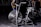 A stationary exercise bike with large fan blades is situated in a dimly lit gym environment. The bike is black with metallic accents and has dual handlebars and a comfortable saddle. Other gym equipment can be seen vaguely in the background, while a person appears to be exercising behind the bike.