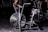 A stationary exercise bike with large fan blades is situated in a dimly lit gym environment. The bike is black with metallic accents and has dual handlebars and a comfortable saddle. Other gym equipment can be seen vaguely in the background, while a person appears to be exercising behind the bike.