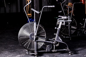 A stationary exercise bike with large fan blades is situated in a dimly lit gym environment. The bike is black with metallic accents and has dual handlebars and a comfortable saddle. Other gym equipment can be seen vaguely in the background, while a person appears to be exercising behind the bike.
