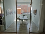 A modern, well-lit conference room with a long table surrounded by green chairs. The walls are adorned with framed artwork, and a large window provides ample natural light. There's a flat-screen TV mounted on the wall alongside minimalist decor.