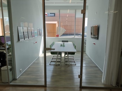 A modern, well-lit conference room with a long table surrounded by green chairs. The walls are adorned with framed artwork, and a large window provides ample natural light. There's a flat-screen TV mounted on the wall alongside minimalist decor.