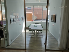 A modern, well-lit conference room with a long table surrounded by green chairs. The walls are adorned with framed artwork, and a large window provides ample natural light. There's a flat-screen TV mounted on the wall alongside minimalist decor.