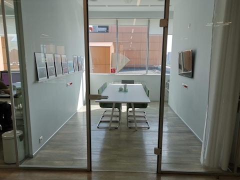 A modern, well-lit conference room with a long table surrounded by green chairs. The walls are adorned with framed artwork, and a large window provides ample natural light. There's a flat-screen TV mounted on the wall alongside minimalist decor.