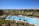 A luxurious resort area featuring a large, serene swimming pool surrounded by numerous palm trees. The pool is shaped uniquely with a round section on one end and an oval pool nearby. Well-maintained lawns and garden areas with tropical vegetation are visible. In the distance, there are landscaped golf courses and a picturesque view of the ocean horizon under a clear blue sky. Modern, colorful buildings are seen in the background.