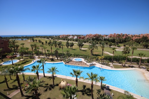 A luxurious resort area featuring a large, serene swimming pool surrounded by numerous palm trees. The pool is shaped uniquely with a round section on one end and an oval pool nearby. Well-maintained lawns and garden areas with tropical vegetation are visible. In the distance, there are landscaped golf courses and a picturesque view of the ocean horizon under a clear blue sky. Modern, colorful buildings are seen in the background.