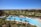 A luxurious resort area featuring a large, serene swimming pool surrounded by numerous palm trees. The pool is shaped uniquely with a round section on one end and an oval pool nearby. Well-maintained lawns and garden areas with tropical vegetation are visible. In the distance, there are landscaped golf courses and a picturesque view of the ocean horizon under a clear blue sky. Modern, colorful buildings are seen in the background.