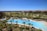 A luxurious resort area featuring a large, serene swimming pool surrounded by numerous palm trees. The pool is shaped uniquely with a round section on one end and an oval pool nearby. Well-maintained lawns and garden areas with tropical vegetation are visible. In the distance, there are landscaped golf courses and a picturesque view of the ocean horizon under a clear blue sky. Modern, colorful buildings are seen in the background.