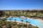 A luxurious resort area featuring a large, serene swimming pool surrounded by numerous palm trees. The pool is shaped uniquely with a round section on one end and an oval pool nearby. Well-maintained lawns and garden areas with tropical vegetation are visible. In the distance, there are landscaped golf courses and a picturesque view of the ocean horizon under a clear blue sky. Modern, colorful buildings are seen in the background.