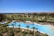 A luxurious resort area featuring a large, serene swimming pool surrounded by numerous palm trees. The pool is shaped uniquely with a round section on one end and an oval pool nearby. Well-maintained lawns and garden areas with tropical vegetation are visible. In the distance, there are landscaped golf courses and a picturesque view of the ocean horizon under a clear blue sky. Modern, colorful buildings are seen in the background.
